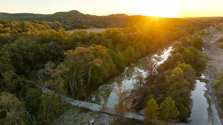 Aerial view of a winding river through a forested valley at golden sunset, with a low stone ford and rolling hills
