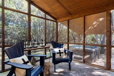Cozy screened wooden porch with vaulted cedar ceiling, two navy Adirondack chairs with cross-pattern pillows, lanterns on a table, and a hot tub on the adjoining deck overlooking a wooded backyard.