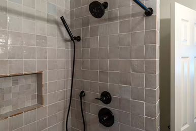 Modern bathroom walk-in shower with glossy gray square tiles, matte black rainfall showerhead, handheld wand, round controls and recessed shelf niche