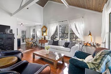 Cozy sunlit farmhouse-style living room with vaulted wood ceiling, hardwood floors, large windows dressed in sheer white curtains, white sofa, leather chairs, rustic coffee table and a small dining nook.