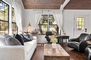 Cozy sunlit modern farmhouse living room with wood-plank ceiling, large black-framed windows and white curtains, white textured sofa with patterned pillows, black leather chairs, rustic wooden coffee table and hardwood floors.