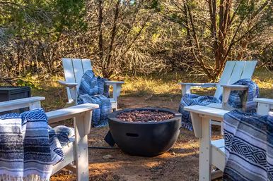 Cozy outdoor fire pit seating in a wooded backyard with white Adirondack chairs draped in blue patterned blankets around a round black fire bowl
