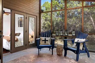 Cozy screened-in porch with two blue Adirondack chairs, cross-pillows, rustic stump side table, lanterns and French doors opening to a bedroom, overlooking a wooded backyard