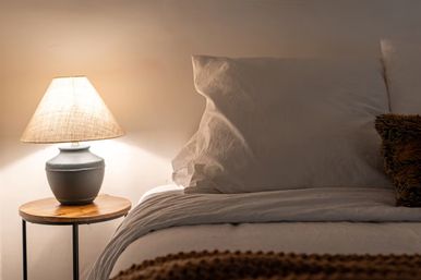 Cozy bedroom with warm-lit bedside lamp on a wooden side table, soft linen pillows and neutral-toned blankets.