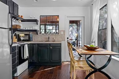 Sunlit cozy eat-in kitchen with dark gray cabinets, white appliances, hardwood floors, a wooden table with metal legs and two chairs, a bowl of green apples, windows with white curtains and a glass backdoor to a porch.