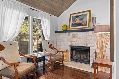 Cozy rustic living room with stone fireplace and framed painting on the mantel, two wooden armchairs with cowhide upholstery, dark hardwood floors, large window with white curtains, vaulted wood ceiling and floor lamp
