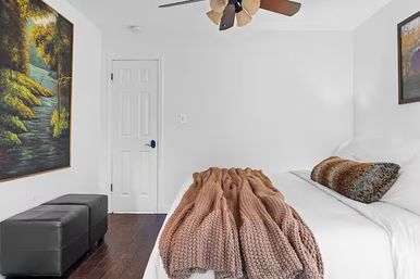 Cozy modern bedroom interior with white walls and dark hardwood floors, bed topped with chunky tan knit throw and faux fur pillow, ceiling fan, black ottomans and framed landscape painting.