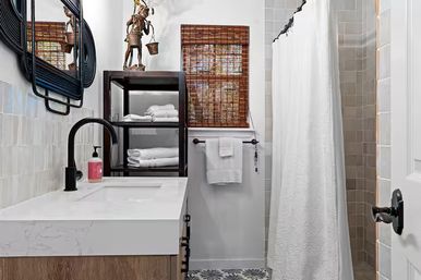 Bright modern small bathroom with white marble countertop vanity, matte black faucet, open wooden shelving with folded towels, woven bamboo window shade, patterned floor tile and neutral tiled shower with white curtain