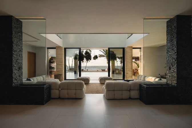 Beachfront modern living room with low tufted white sofas and stone pillars framing floor-to-ceiling glass opening to an infinity pool, palm trees, and ocean view
