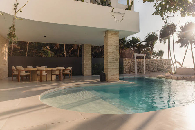 Sunlit modern villa patio with a curved turquoise pool, shaded outdoor dining area with wooden chairs, stone columns and swaying palm trees framing an ocean beach view