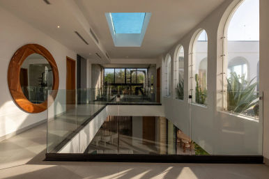 Bright modern hallway with skylight, glass balustrade overlooking a lower level, round wooden mirror on the wall, and a row of arched windows planted with succulents and palm views
