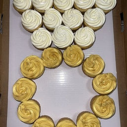 Swirled white and gold buttercream cupcakes arranged in a decorative ring on a white board inside a bakery box — elegant party cupcakes for celebration or event