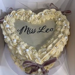 Heart-shaped cream buttercream cake in a box with piped rosette border, edible pearl sprinkles, black cursive message on top and a small velvet bow accent.