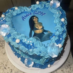 Heart-shaped blue celebration cake with edible photo of a woman and "Aries SZN" text, piped turquoise and navy buttercream swirls, pearl and crystal-like sugar decorations