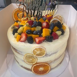 Layered naked-style birthday cake with thin white buttercream, piled with fresh mixed fruit (strawberries, blueberries, blackberries, mango, kiwi), a silver Happy Birthday mirror topper, and dried citrus slice accents on the side.