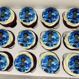 Box of a dozen red velvet cupcakes with white swirled frosting and round blue edible toppers showing a cartoon boy over a blue camouflage pattern, arranged in neat rows.