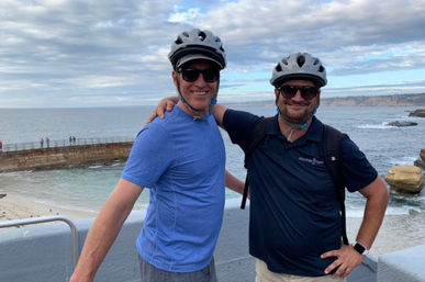 Two people wearing bike helmets and sunglasses smiling with arms around each other on an oceanfront overlook, rocky shoreline and boardwalk visible under a cloudy sky