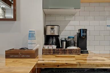 Compact coffee station on a polished wood counter with a drip coffee maker, single-serve brewer, stacked paper cups, jar of beans and woven tray against a white subway tile backsplash.