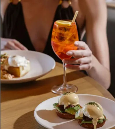 Brunch at a cafe: two avocado eggs Benedict on toasted muffins on a white plate, with a bright orange aperitif garnished with lemon held by a diner.