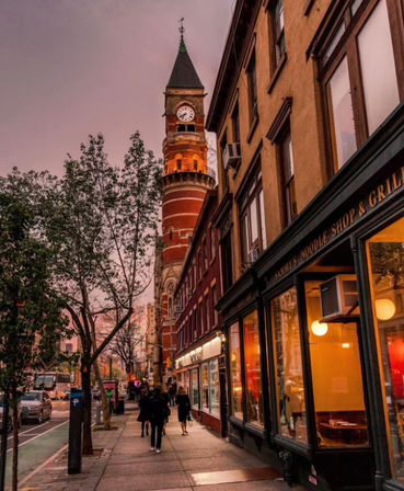 Downtown city street at dusk with a historic red-brick clock tower, tree-lined sidewalk, pedestrians, and warm-lit storefront and restaurant windows.