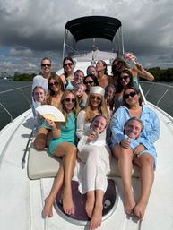 Yacht party: a group of women in summer outfits and sunglasses lounging on a luxury boat bow, smiling and holding cardboard cutouts of a man’s face with coastal water and a cloudy sky in the background