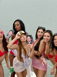 Bachelorette boat party: group of women in swimsuits on a boat deck posing with printed face cutouts; bride-to-be wearing a sash and captain hat, water in the background.