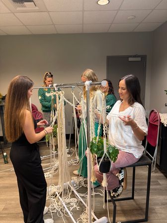 Women at an indoor macramé workshop crafting plant hangers from cotton cords on a metal rack with a potted plant.