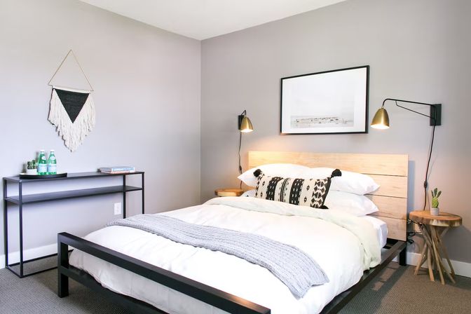 Bright modern minimalist bedroom with queen bed, light wood headboard and black metal frame, neutral gray walls, patterned throw pillow, wall sconces, macramé wall hanging, console table and small potted plant
