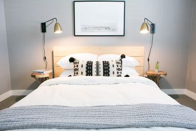 Cozy modern bedroom interior with white bedding, gray knit throw, black-and-white patterned lumbar pillow with pom-poms, light wood headboard, brass wall sconces, round wooden side tables with books and a small succulent, and framed art on a gray wall.
