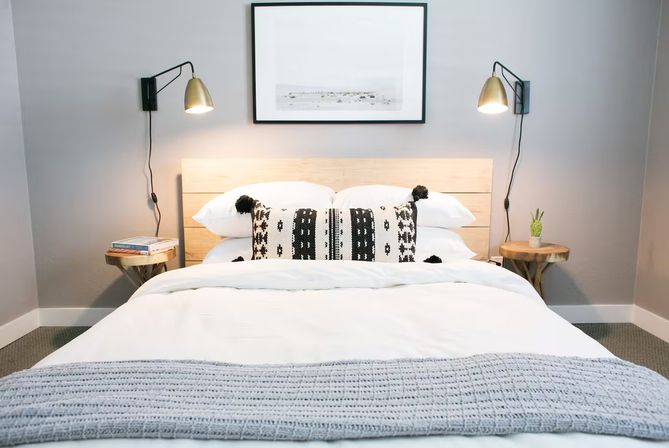 Cozy modern bedroom interior with white bedding, gray knit throw, black-and-white patterned lumbar pillow with pom-poms, light wood headboard, brass wall sconces, round wooden side tables with books and a small succulent, and framed art on a gray wall.