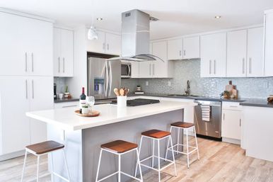 Bright modern white kitchen with a large island, four wooden-top bar stools, stainless steel appliances and range hood, mosaic tile backsplash and light wood-look flooring.