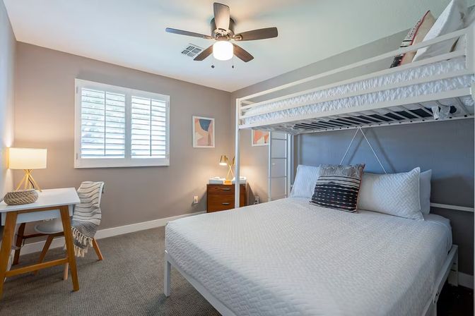 Cozy modern bedroom with white bunk bed (full over twin), neutral gray walls, ceiling fan, desk, nightstand and shuttered window