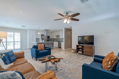 Bright open-concept living room and kitchen with brown leather sofa, blue armchairs, live-edge wooden coffee table, patterned rug, ceiling fan, TV on a wooden console, and a white kitchen island with bar stools.
