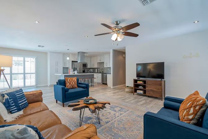 Bright open-concept living room and kitchen with brown leather sofa, blue armchairs, live-edge wooden coffee table, patterned rug, ceiling fan, TV on a wooden console, and a white kitchen island with bar stools.