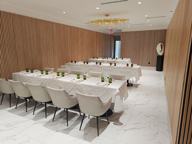Modern private dining room set for a meal with long white-tablecloth banquet tables, beige upholstered chairs, green glassware, vertical wood-slat walls, marble tile floor and a gold branch chandelier.