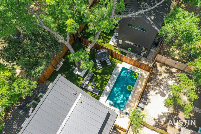 Aerial view of a modern backyard: narrow rectangular pool with two yellow float rings, sun-dappled artificial turf, lounge chairs under mature trees and a wooden fence.