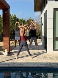 Sunny outdoor fitness class on a modern home's poolside patio — a group of women following an instructor doing arms-up stretches with trees and backyard lawn in the background.