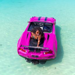 Person in sunglasses posing playfully on a hot-pink sports-car-style boat floating in crystal-clear turquoise shallow tropical water — vibrant beach vacation vibe.