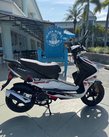 Black-and-white motor scooter with red accents parked in front of a large blue Adirondack chair with a pineapple logo, outside a palm-lined tropical outdoor plaza with café seating on a sunny day.