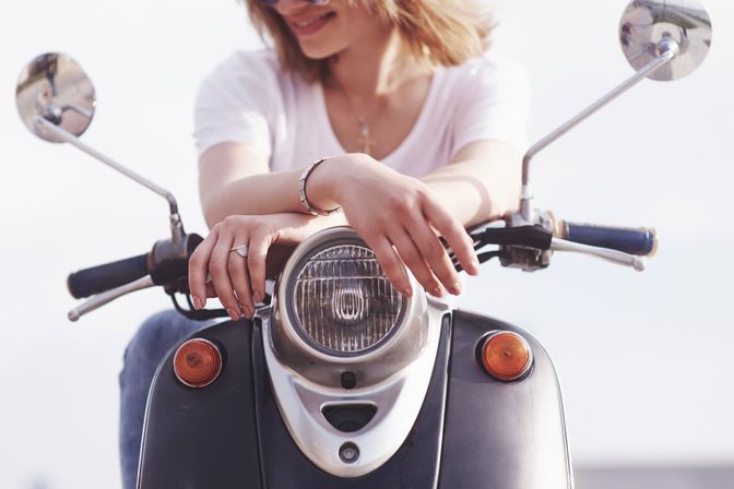 Close-up of a smiling person leaning on a vintage scooter, hands draped over the round headlight and handlebars on a sunny outdoor ride.