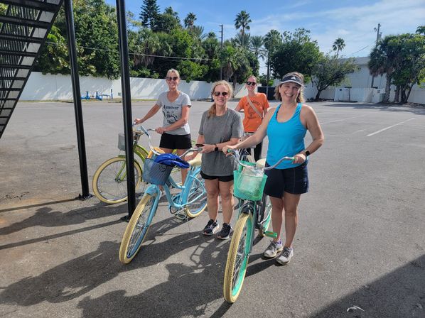 Four women smiling and ready to ride colorful cruiser bikes in a sunny beach‑town parking lot with palm trees and clear blue sky.