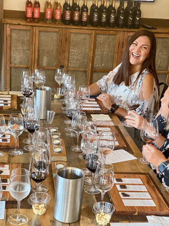 Smiling woman at a rustic winery tasting room enjoying a wine tasting; long wooden table set with multiple wine glasses, wine flight cards, small snack cups and bottles on a back shelf.