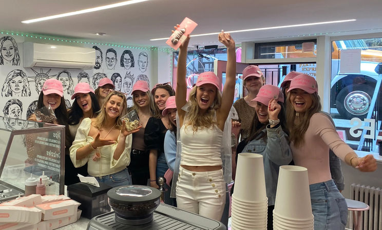 Energetic group of friends wearing pink caps cheering and holding cookies inside a pink‑themed city dessert shop by a counter with takeaway cups and an illustrated wall mural.