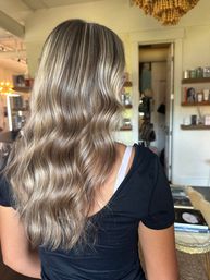 Back view of person with glossy ash-blonde balayage and soft beachy waves, styled shoulder-length hair in a bright hair salon setting.