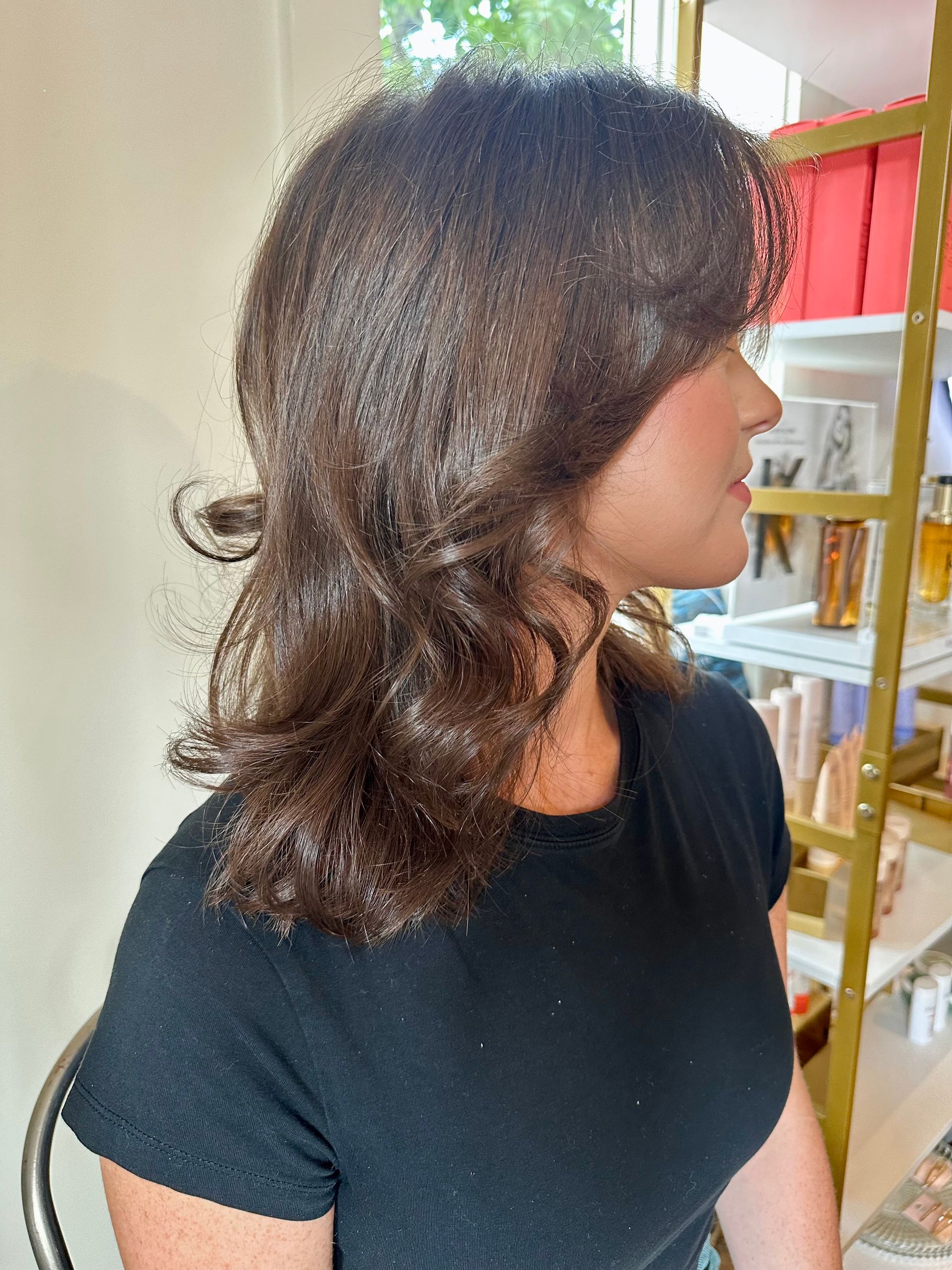 Shoulder-length glossy wavy brown haircut with soft curtain bangs, styled in a bright salon setting with product shelves in the background.