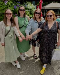 Four friends linking pinkies and smiling on a sunny outdoor garden patio with purple hydrangeas, wearing summer dresses, sunglasses and casual shoes.