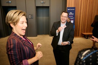 Man in a suit showing a playing card and making a woman laugh during a corporate networking event in a conference lobby with elevators and wood-paneled walls.