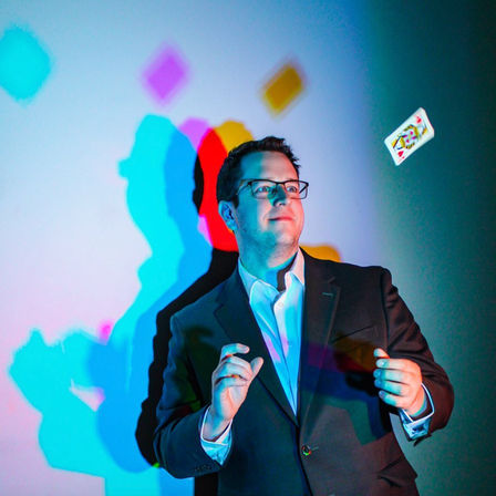 Man in a suit and glasses tossing a playing card in front of a wall lit with colorful cyan, magenta and yellow layered shadows — playful card trick portrait.