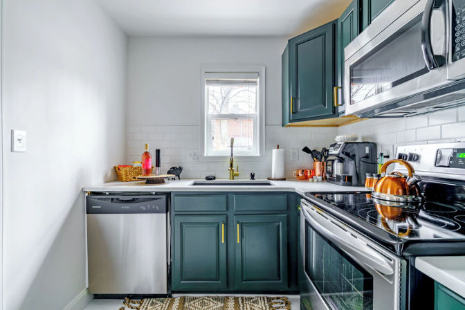 Bright compact modern kitchen with dark green cabinets and brass pulls, white subway tile backsplash, stainless steel appliances, gold faucet under a small window, countertop coffee maker, basketed bottle, and a shiny copper kettle on the stove.