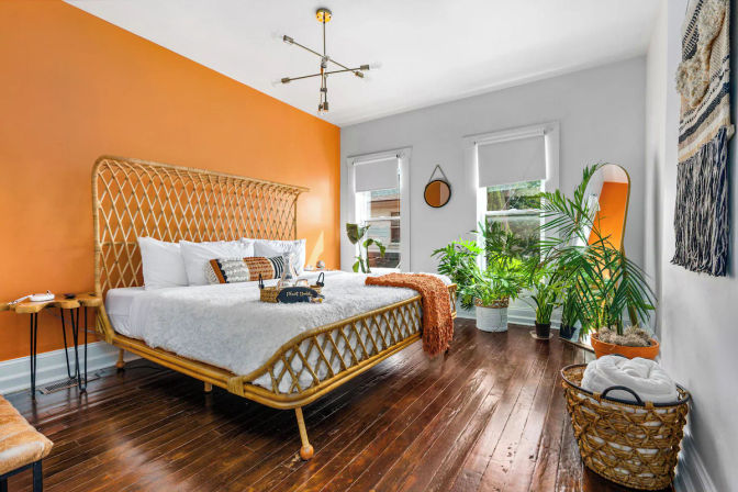 Cozy sunlit bohemian bedroom with woven rattan bed, orange accent wall, dark hardwood floors, white bedding, brass chandelier, full-length mirror and lush indoor plants.
